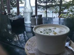 A cup of clam chowder on a dark table by a window looking out to the water
