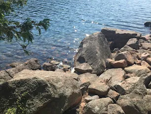 Clear water over pink granite cobbles at the edge of Jordan Pond with wooded hills beyond