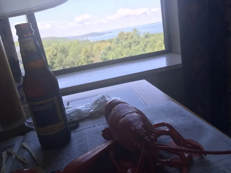 A whole steamed lobster and a bottle of Atlantic blueberry ale on newspaper at a window over Frenchman Bay