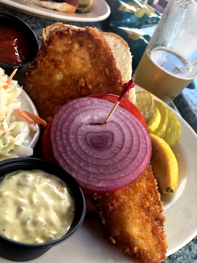 A breaded fish sandwich on white bread with coleslaw, pickles, and a beer at a Cape May lunch spot