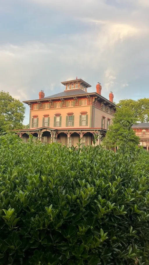 A tall salmon and green Victorian cottage with a mansard tower and a wide porch rising behind a clipped hedge in Cape May