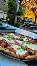 A wood-fired margherita pizza on a metal tray at the Kaatskeller, picnic tables and autumn trees behind it