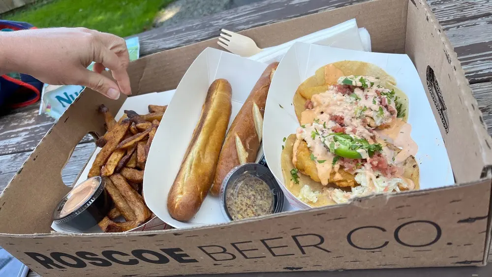 A cardboard box stamped Roscoe Beer Co holding fish tacos, fries, and soft pretzel rolls on a picnic table