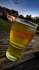 A glass of pale beer marked Russian Mule Brewery on a picnic table at dusk, the brewery building lit behind it