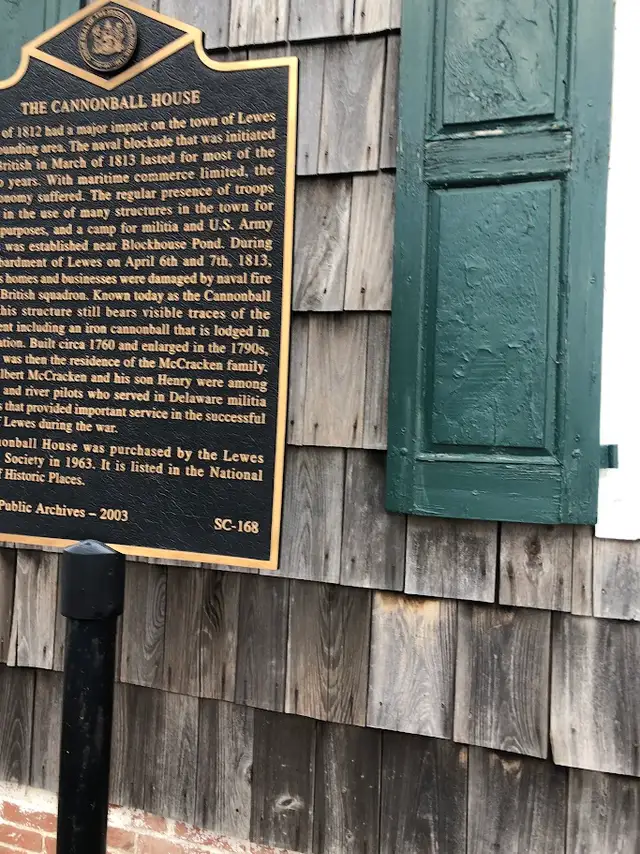 A bronze historical marker for the Cannonball House mounted on a cedar-shingled wall beside a dark green shutter in Lewes