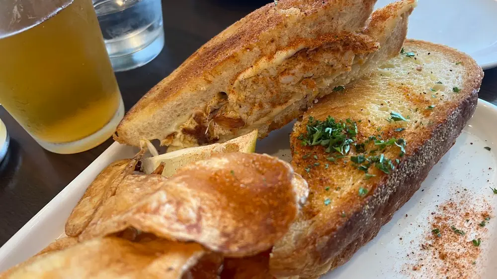 A grilled crab melt cut in half on a white plate with house chips and a beer at Crooked Hammock Brewery in Lewes
