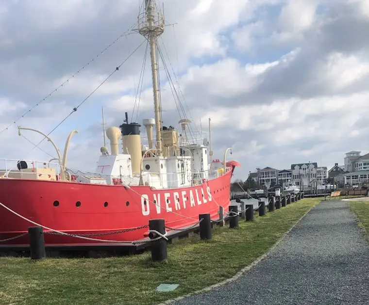 The bright red hull of the lightship Overfalls moored on the grass beside the gravel path at Lewes Canalfront Park