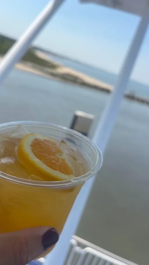 A plastic cup of orange crush held up on a ferry deck with the dunes of Cape Henlopen and the breakwater behind it