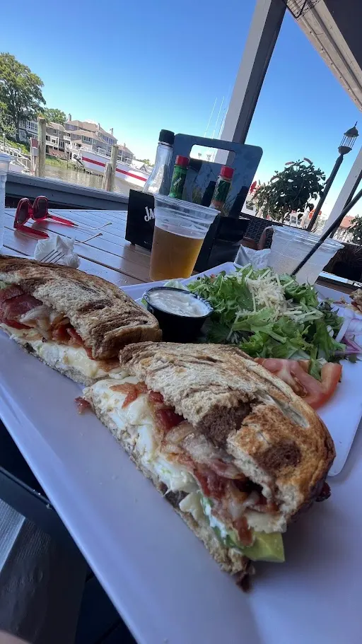 A grilled crab and bacon sandwich with a salad and a beer on a white table, the Lewes canal and boats visible behind