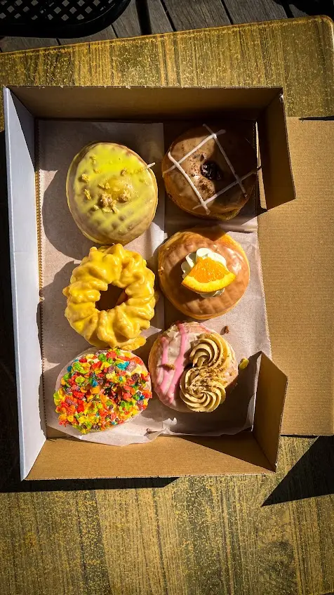 An open box of six elaborately decorated Deviant Donuts on a sunlit picnic table in Olde Mistick Village