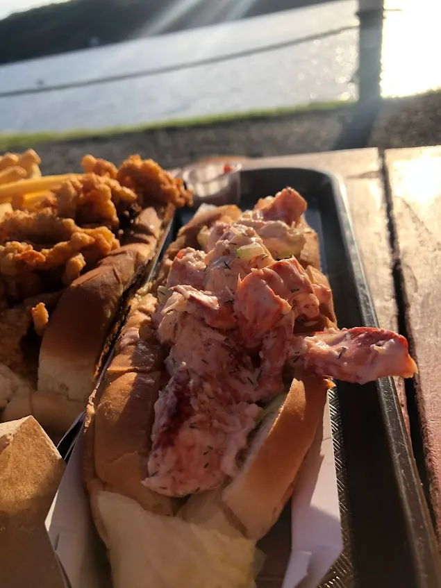 An overstuffed cold lobster roll with a side of fried clams on a tray, the water and evening light behind it