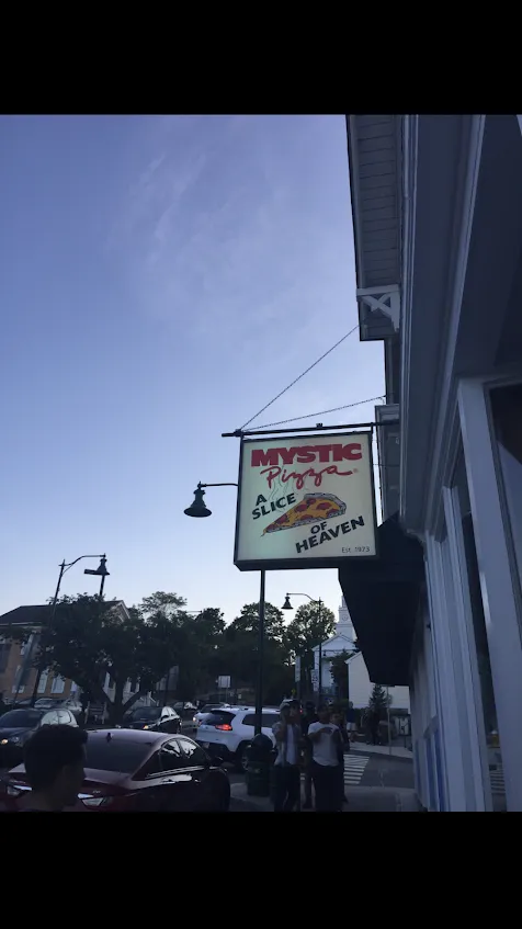 The hanging sign for Mystic Pizza reading A Slice of Heaven, Est. 1973, against an evening sky