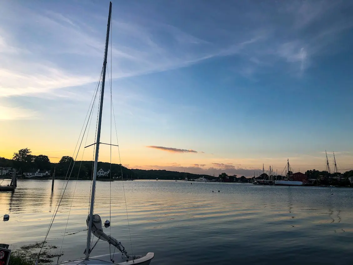The Mystic River at dusk, a moored sailboat in the foreground and the masts of tall ships along the far bank