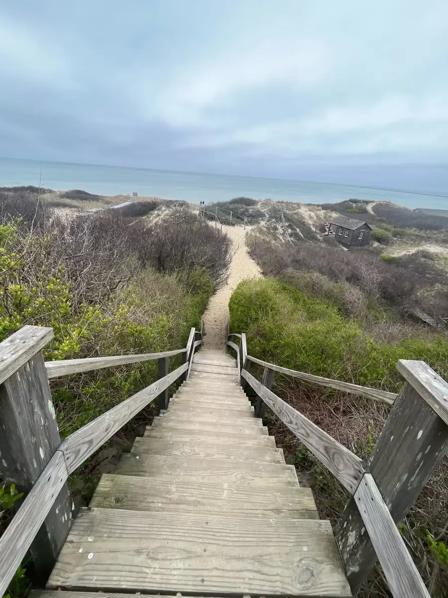 Wooden stairs leading down through dune grass to a Nantucket beach under a grey spring sky, a shingled cottage tucked into the bluff