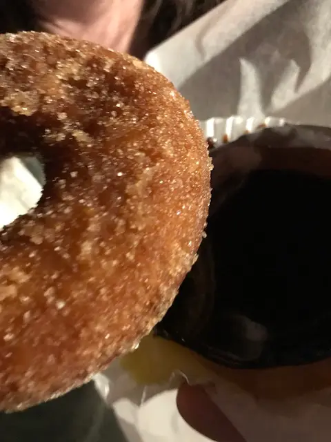A hot cinnamon-sugar cider donut held up next to a cup of coffee in a paper bag at night