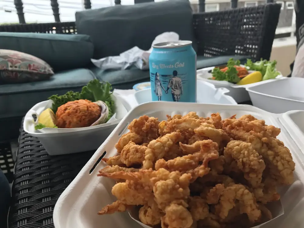 A takeout tray heaped with fried clams and a crab cake, a can of local cider beside it on a porch