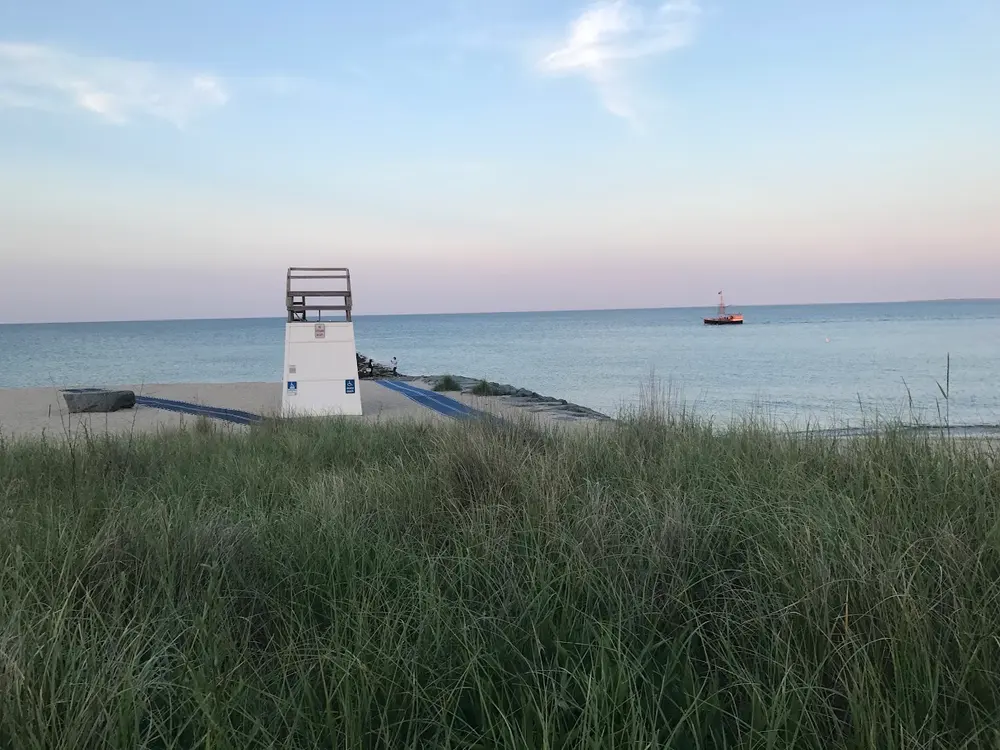 The Oak Bluffs town beach at dusk seen through the dune grass, a lifeguard stand and a schooner offshore