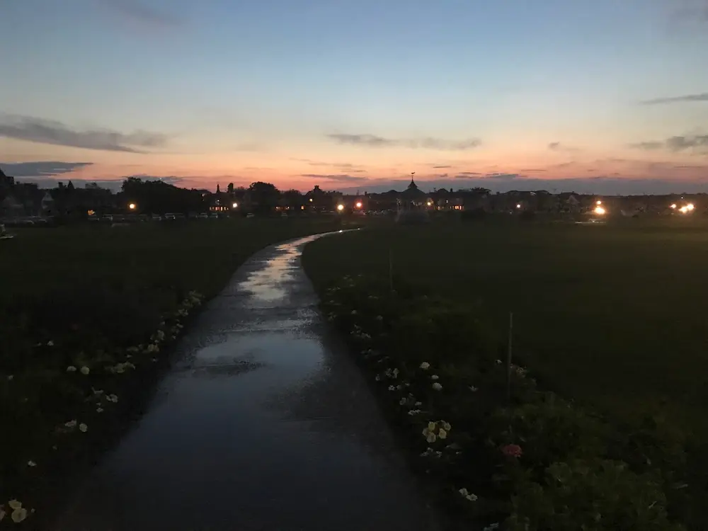 A wet path curving across Ocean Park toward town lights at dusk, the bandstand silhouetted against a pink sky
