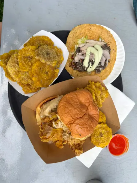An overhead spread on an outdoor table of a fried chicken sandwich in a paper boat, a plate of tostones, and an open taco with sliced chicken and green crema