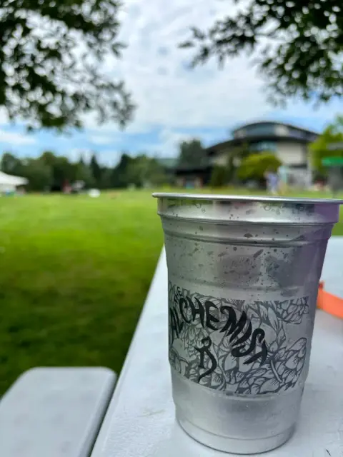 A metal cup printed The Alchemist with hop-vine artwork, beaded with condensation, on a white picnic table on the brewery's green lawn with the curved-roof building behind