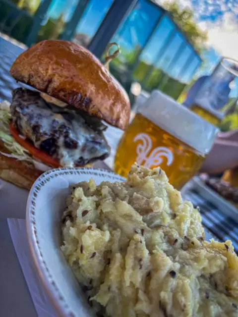 A cheeseburger on a brioche bun with lettuce and tomato next to a bowl of caraway-flecked mashed potatoes with braised cabbage, and a glass of golden von Trapp lager