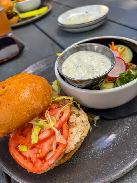 A grilled chicken sandwich with lettuce and tomato beside a small green side salad with radish and cucumber and a cup of herb dressing, on a slate patio table