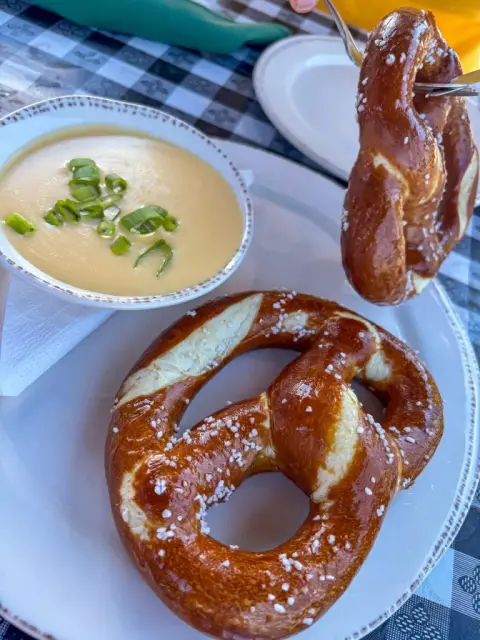 A large golden Bavarian pretzel with pearl salt on a white plate beside a bowl of pale cheddar lager soup topped with scallions, on a blue gingham cloth
